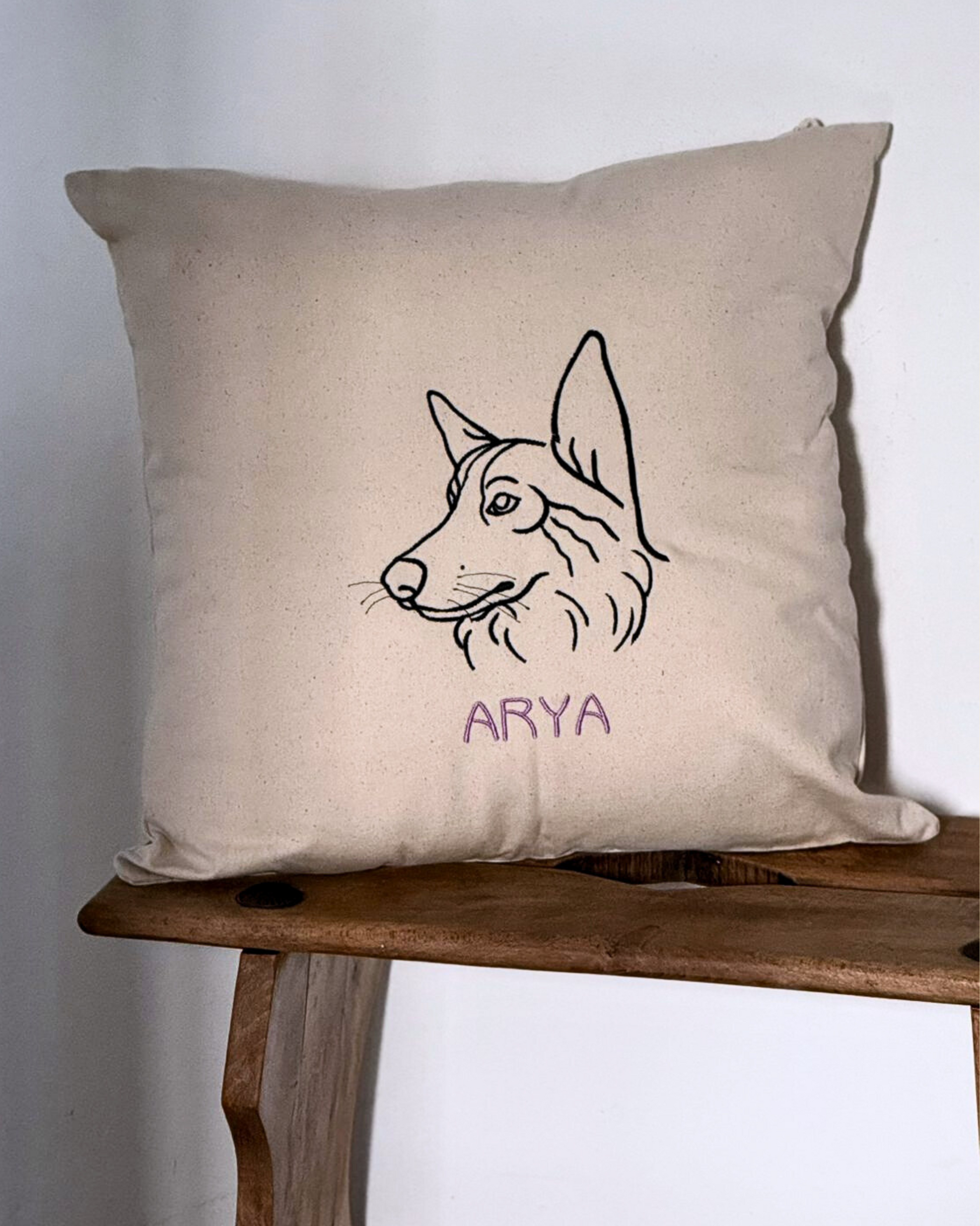 El Cojín personalizado es de algodón beige, con diseño minimalista: línea negra que dibuja la cabeza de un perro y el nombre ARYA bordado en púrpura, colocado sobre un taburete de madera y fondo blanco.