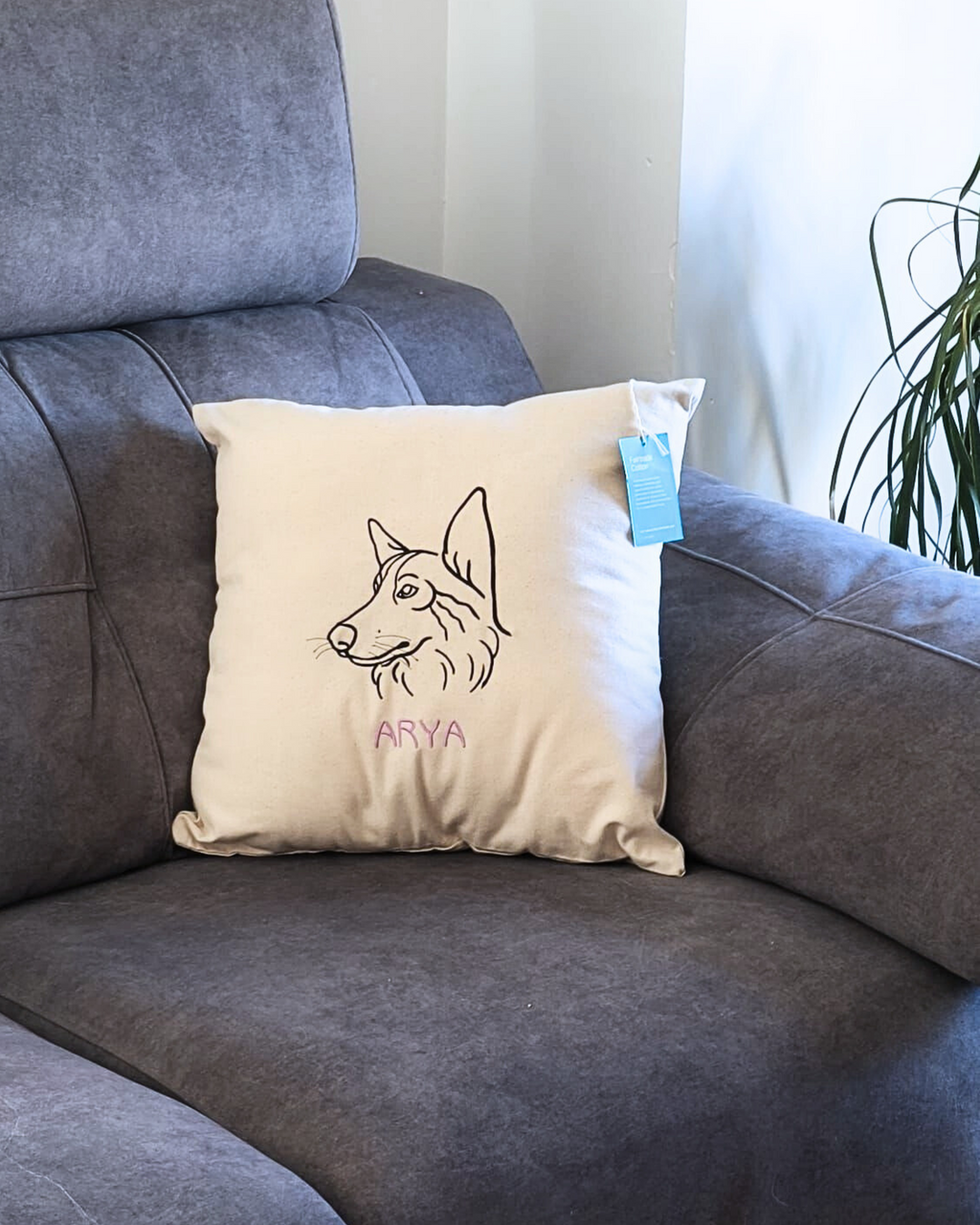 A cream-colored Cojín personalizado with a dog’s head embroidered and the name ARYA in pink sits on a gray armchair. A blue tag is attached to the pillow, with a plant seen in the background.