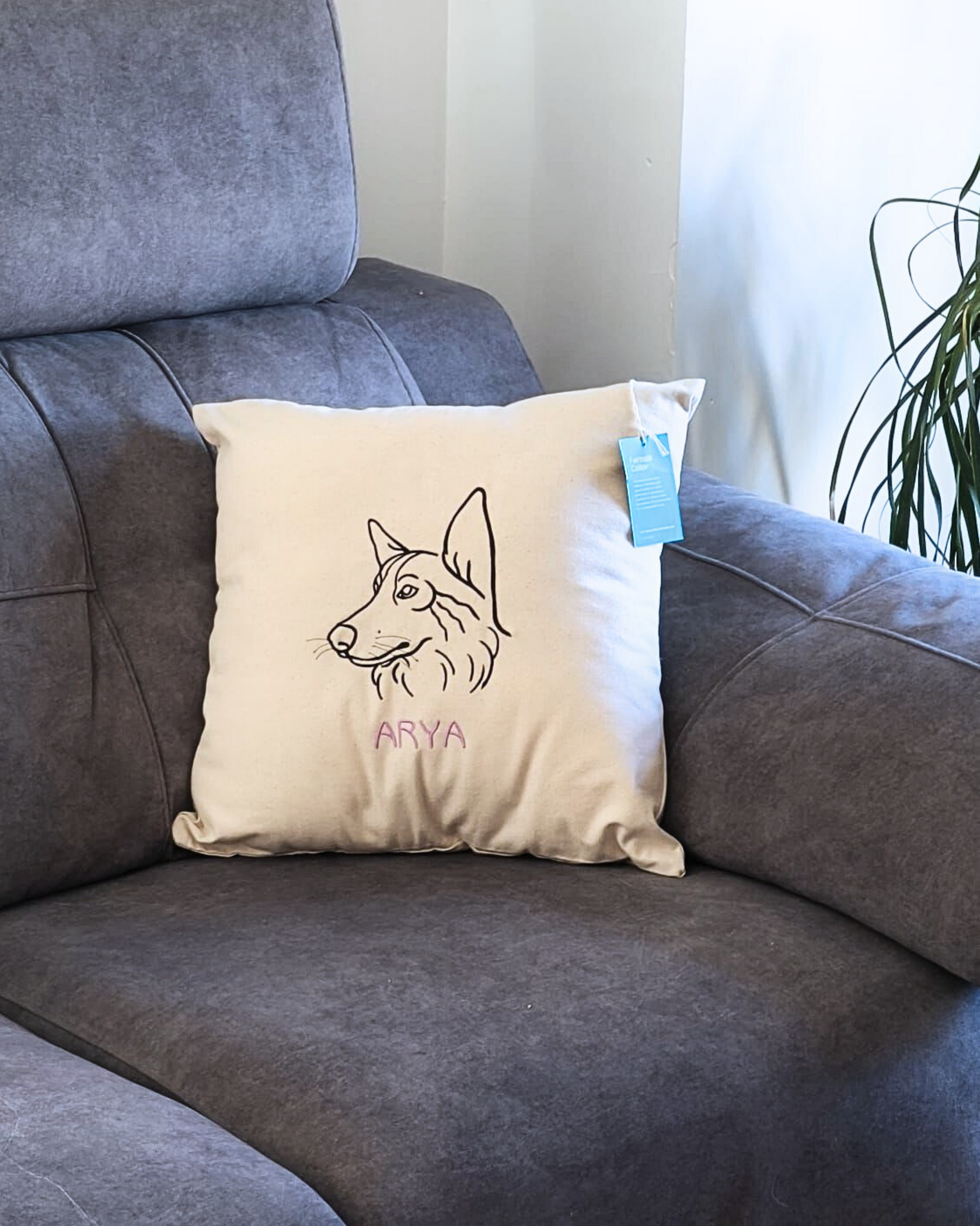 A cream-colored Cojín personalizado with a dog’s head embroidered and the name ARYA in pink sits on a gray armchair. A blue tag is attached to the pillow, with a plant seen in the background.