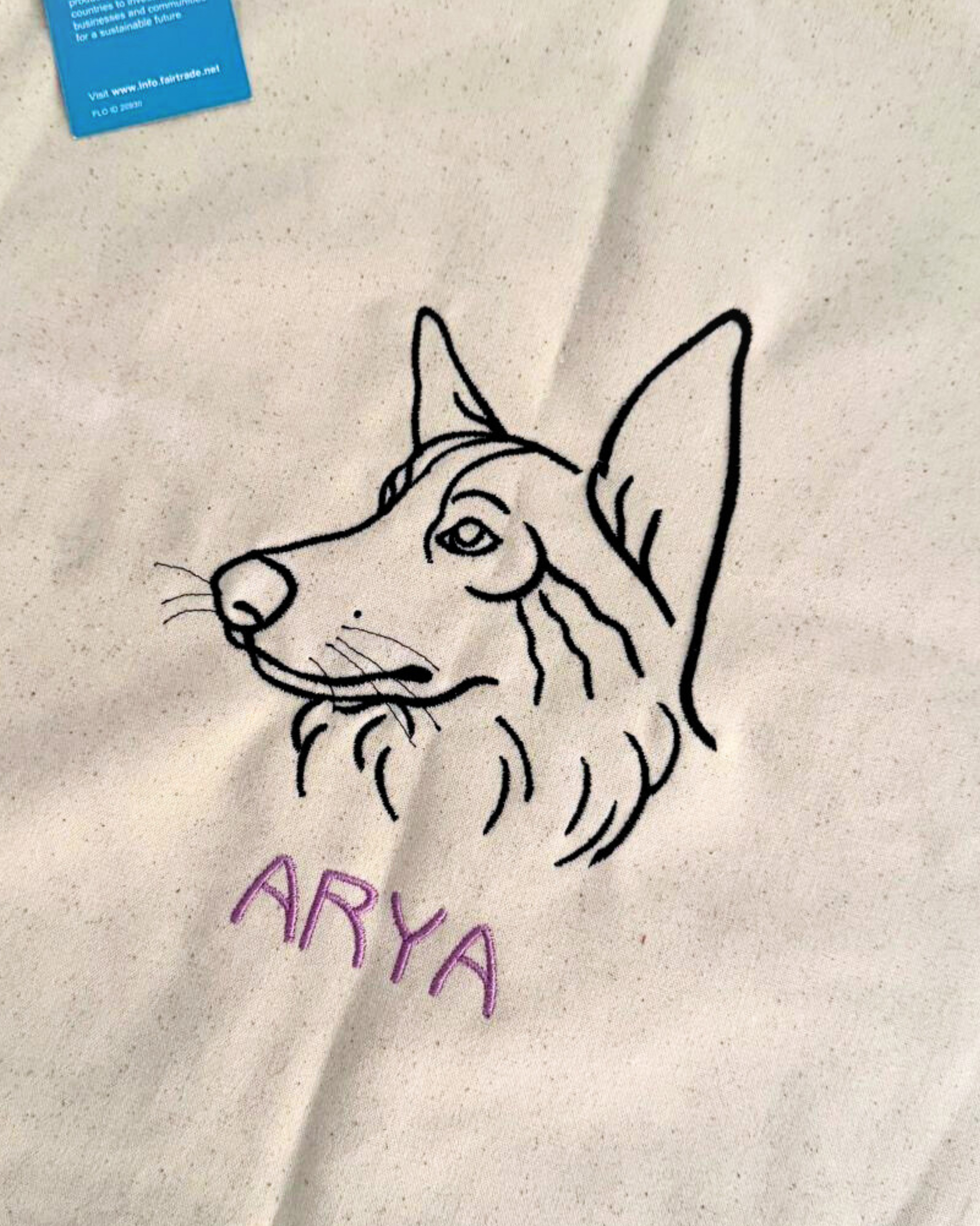 A black line drawing of a dogs head is embroidered on white Cojín personalizado fabric in a minimalistic style, with the name ARYA stitched in purple letters below. A blue card appears partially in the top left corner.